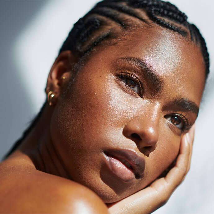 A close-up portrait of a woman with glossy skin and neatly braided cornrows, resting her face in her hand with a calm expression