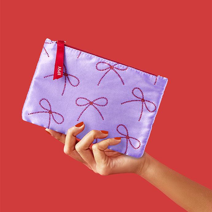 A hand with red nail polish holds a lavender IPSY glam bag decorated with red glitter bows against a red background.