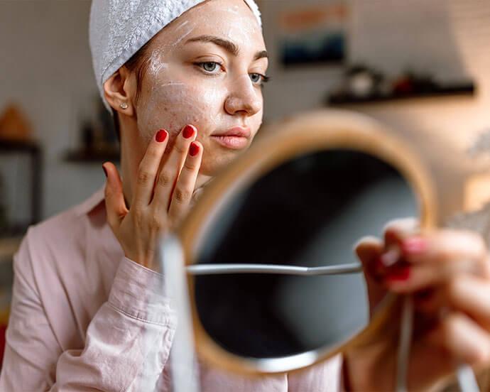 An image of a woman with an acne-prone face is following her skincare routine while holding a mirror, has red-painted fingernails and is dressed in a long-sleeved, baby pink top