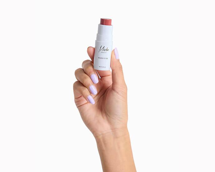 A hand with light purple nail polish is holding a white blush stick with a pink shade exposed