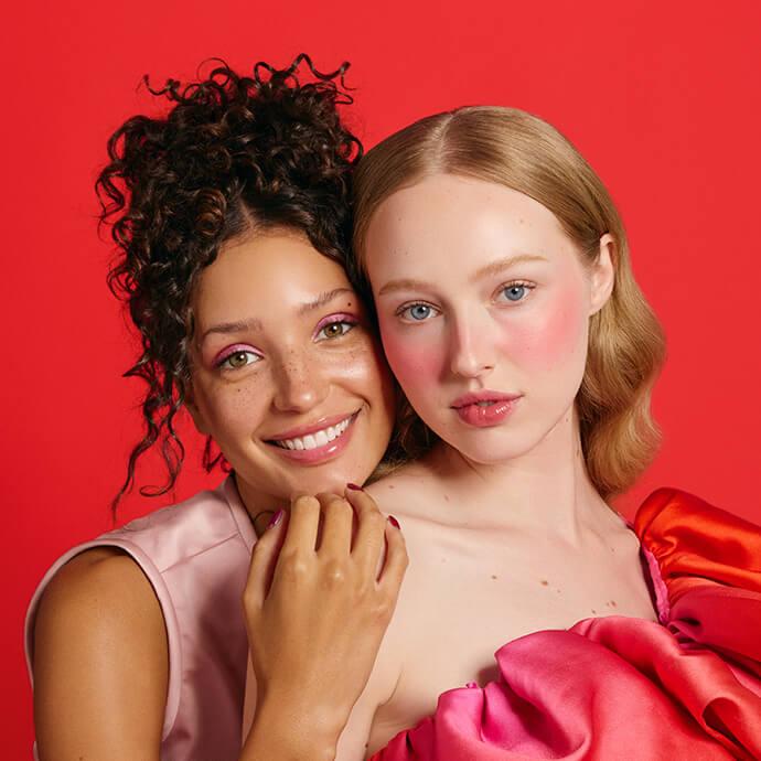 Two people pose closely against a bright red background—one smiling with curly hair and pink eye makeup, the other with wavy blonde hair and bold pink blush—both wearing pink and red fashion looks.