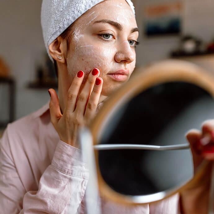 An image of a woman with an acne-prone face is following her skincare routine while holding a mirror, has red-painted fingernails and is dressed in a long-sleeved, baby pink top