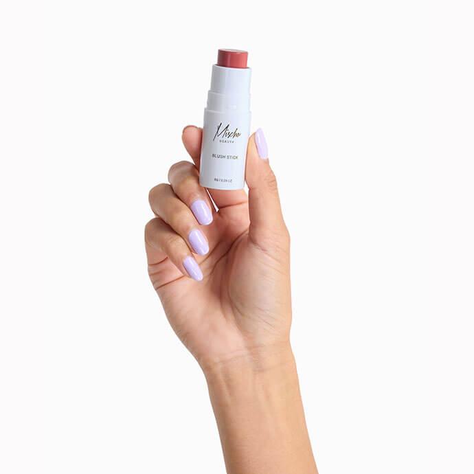 A hand with light purple nail polish is holding a white blush stick with a pink shade exposed