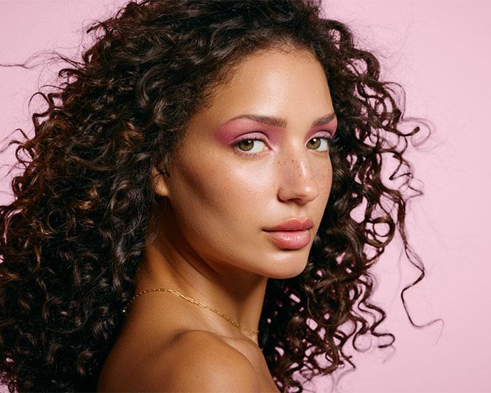 A person with long curly hair poses against a pink background, wearing soft glam makeup with rosy pink eyeshadow, dewy skin, subtle freckles, and glossy lips, accessorized with a delicate gold necklace.