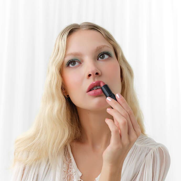 Woman with wavy blonde hair and in a white blouse applies pink lipstick while looking thoughtfully upward