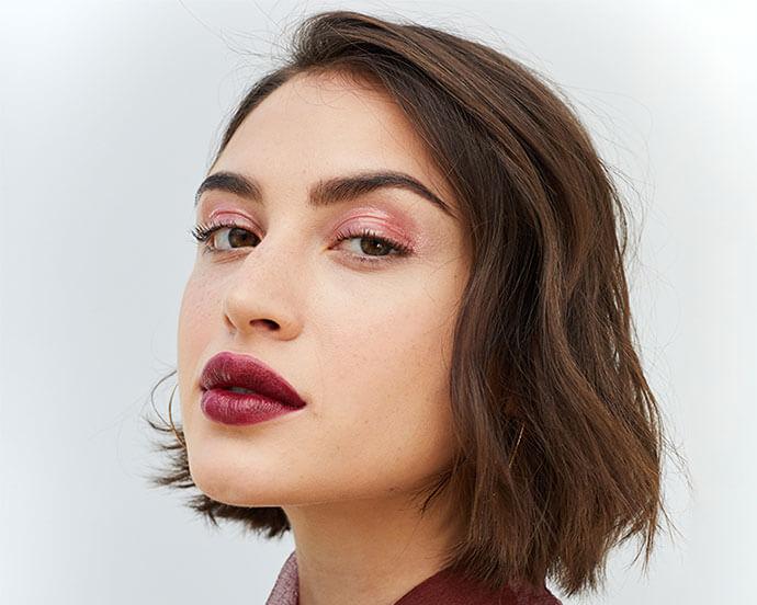 A photo of a woman that has a chic, short brown bob with subtle waves, bold burgundy lipstick, soft pink eye makeup, and a sheer burgundy bow-tied top