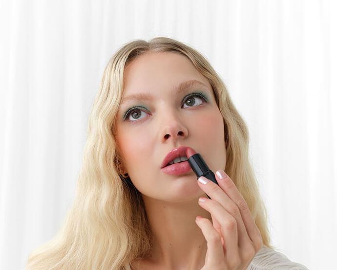 Woman with wavy blonde hair and in a white blouse applies pink lipstick while looking thoughtfully upward