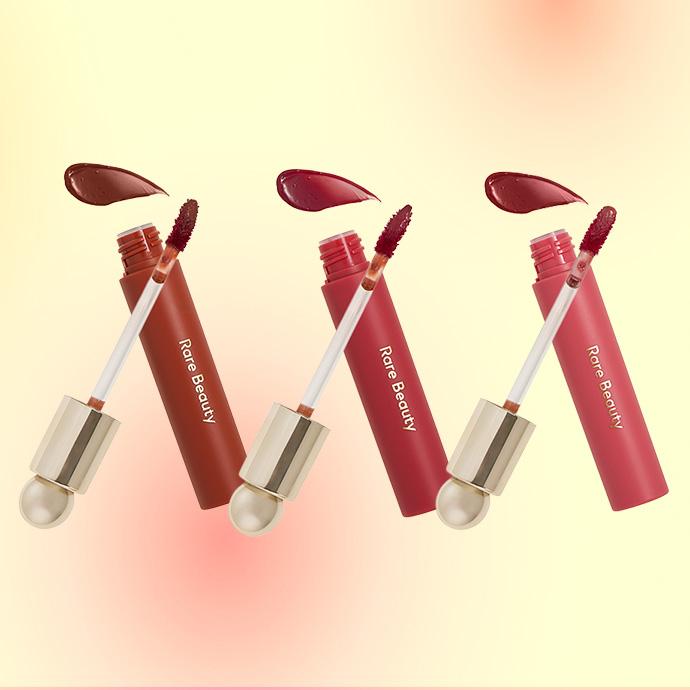 3 RARE BEAUTY Soft Pinch Tinted Lip Oils against an ombre yellow and orange background