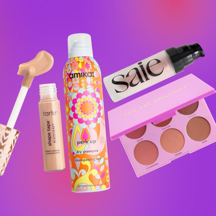 A flat lay of beauty products on a purple-pink gradient background, featuring Tarte Shape Tape Radiant concealer, amika dry shampoo, Saie highlighter, and a cheek face palette.