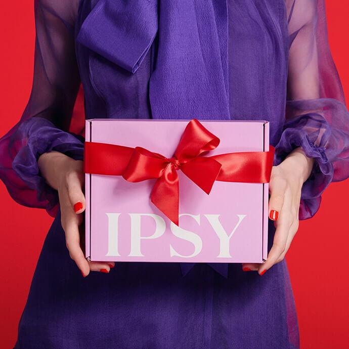 A person wearing a sheer purple blouse holds a pink IPSY gift box tied with a red satin ribbon against a red background.
