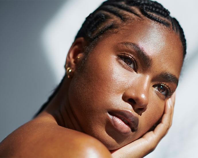A close-up portrait of a woman with glossy skin and neatly braided cornrows, resting her face in her hand with a calm expression