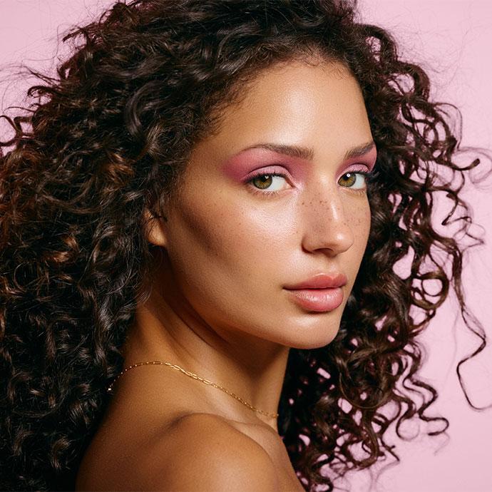 A person with long curly hair poses against a pink background, wearing soft glam makeup with rosy pink eyeshadow, dewy skin, subtle freckles, and glossy lips, accessorized with a delicate gold necklace.