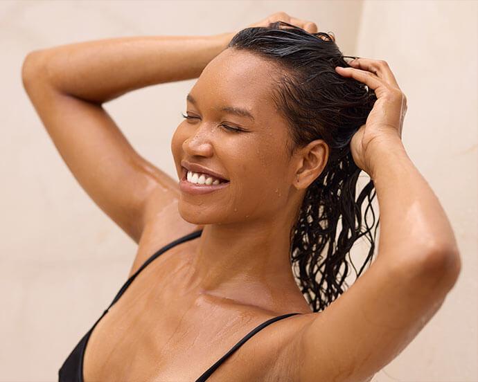 A woman is smiling with her eyes closed while washing her wet hair in the shower