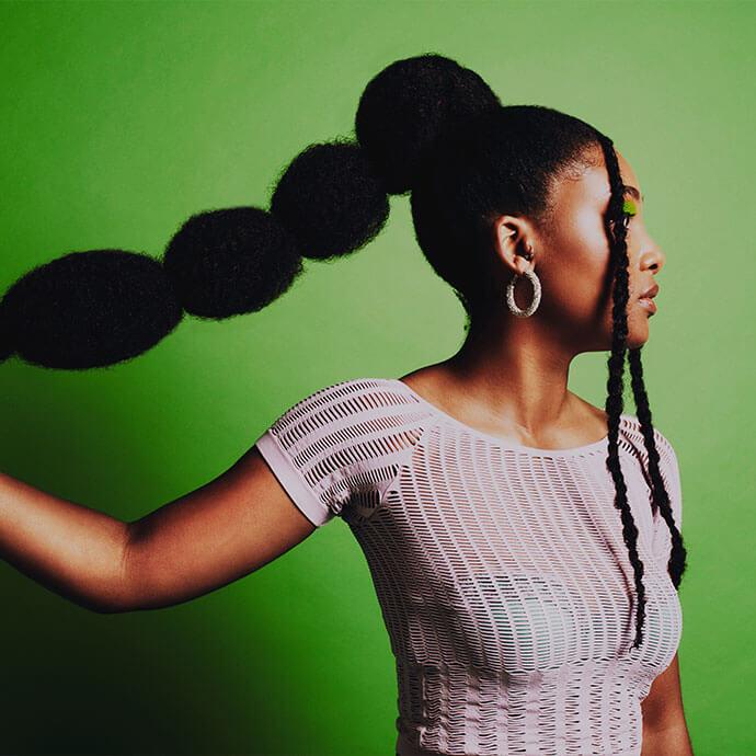 An image of a woman of color with a voluminous braided ponytail against a bright green background, wearing a white textured top and hoop earrings