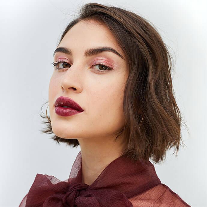 A photo of a woman that has a chic, short brown bob with subtle waves, bold burgundy lipstick, soft pink eye makeup, and a sheer burgundy bow-tied top