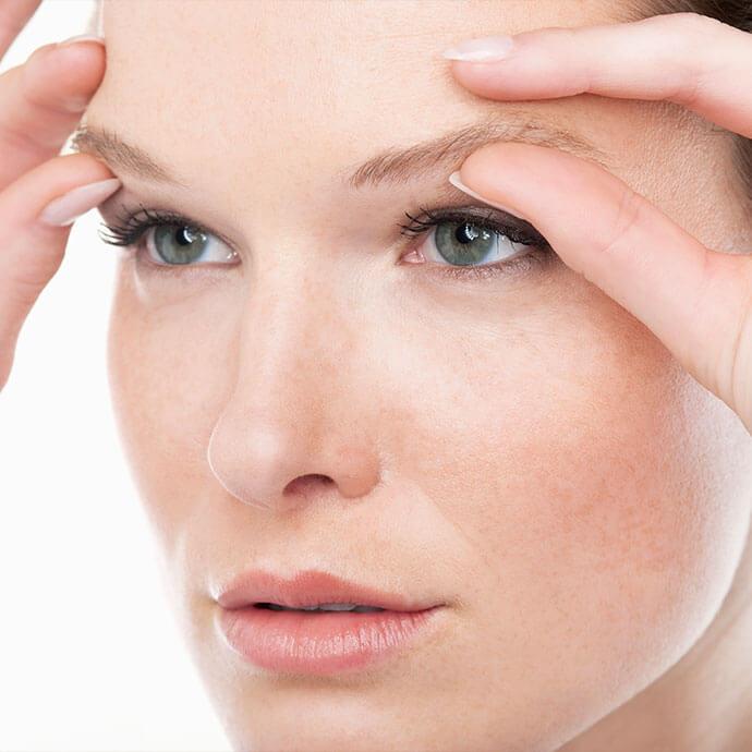 A close-up of a fair-skinned woman with striking blue eyes and light brown eyebrows, softly touching her eyebrows and forehead