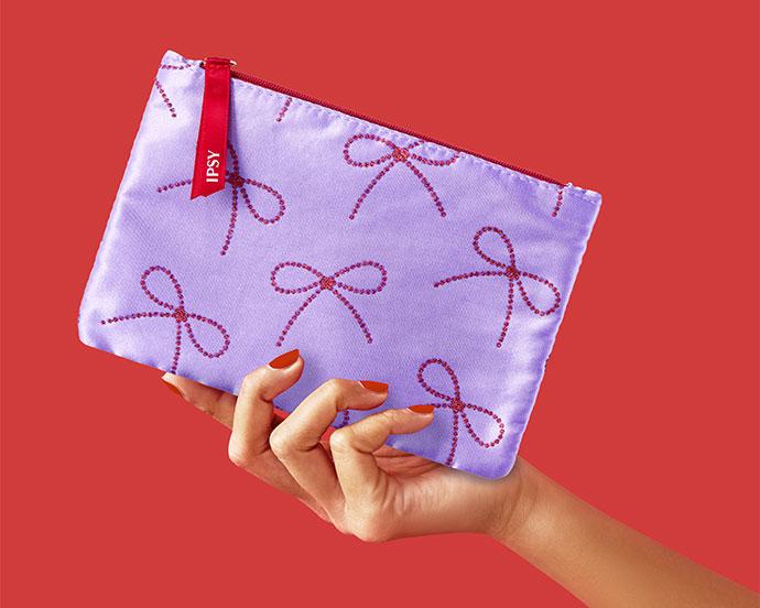 A hand with red nail polish holds a lavender IPSY glam bag decorated with red glitter bows against a red background.