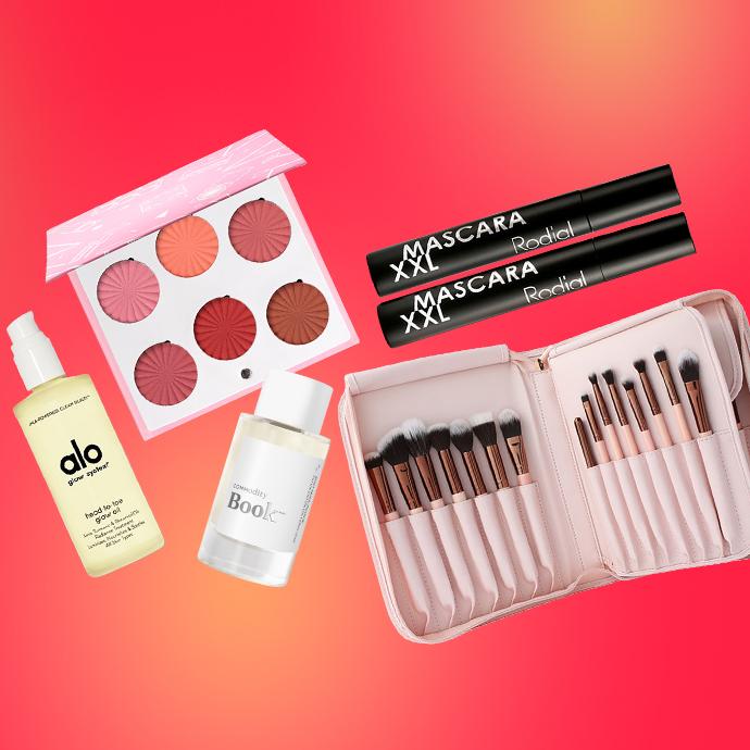 A flat lay of beauty products on a red-to-orange gradient background, featuring Alo Glow System body oil, Commodity Book perfume, Rodial XXL mascara, a blush palette, and a pink brush set in a zip-up case.