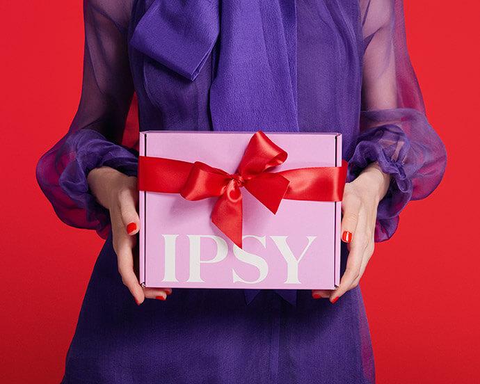 A person wearing a sheer purple blouse holds a pink IPSY gift box tied with a red satin ribbon against a red background.