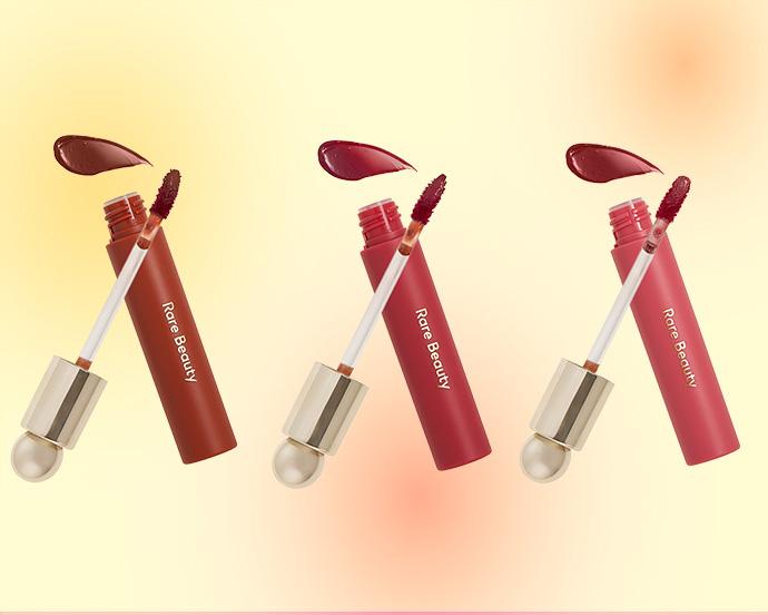 3 RARE BEAUTY Soft Pinch Tinted Lip Oils against an ombre yellow and orange background