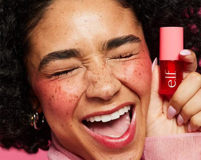 An image of a joyful young woman with curly hair and freckles, flashing a bright smile while holding a pink-lidded pink bottle of ELF lip product against a pink background
