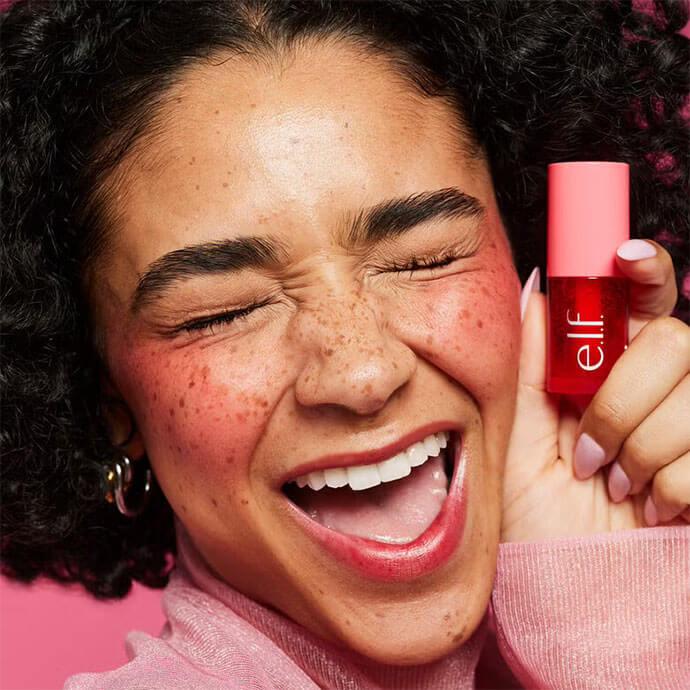 An image of a joyful young woman with curly hair and freckles, flashing a bright smile while holding a pink-lidded pink bottle of ELF lip product against a pink background
