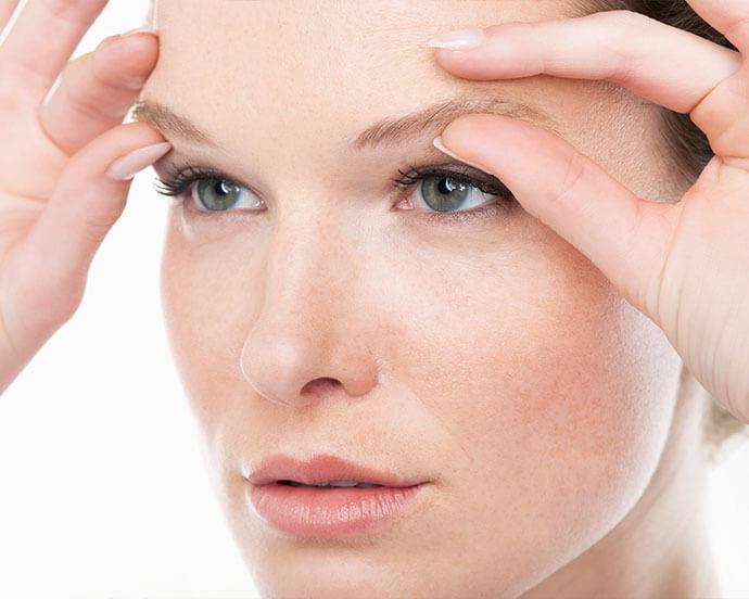 A close-up of a fair-skinned woman with striking blue eyes and light brown eyebrows, softly touching her eyebrows and forehead