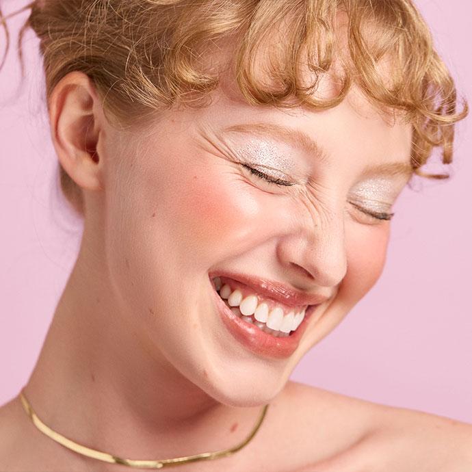 A close-up of a person smiling with eyes closed, wearing shimmery silver eyeshadow, rosy blush, glossy lips, and a gold necklace, against a soft pink background.