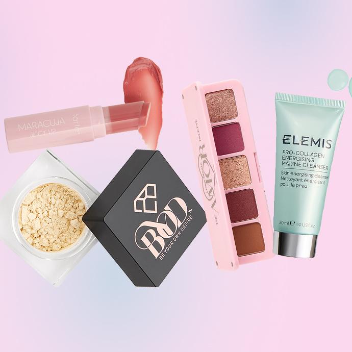 A flat lay of beauty products on a pink-to-blue gradient background, including Tarte Maracuja Juicy Lip, loose setting powder, BYOD compact, a LUNA MAGIC eyeshadow palette, and an ELEMIS cleanser.