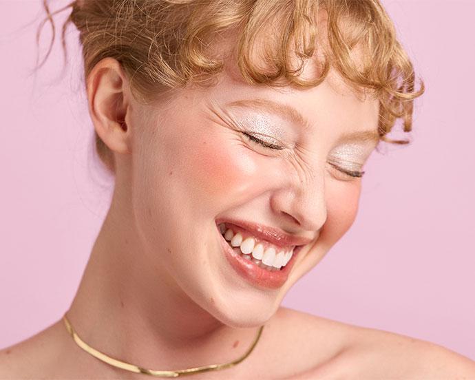 A close-up of a person smiling with eyes closed, wearing shimmery silver eyeshadow, rosy blush, glossy lips, and a gold necklace, against a soft pink background.