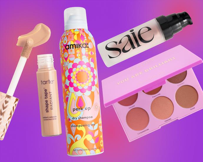 A flat lay of beauty products on a purple-pink gradient background, featuring Tarte Shape Tape Radiant concealer, amika dry shampoo, Saie highlighter, and a cheek face palette.