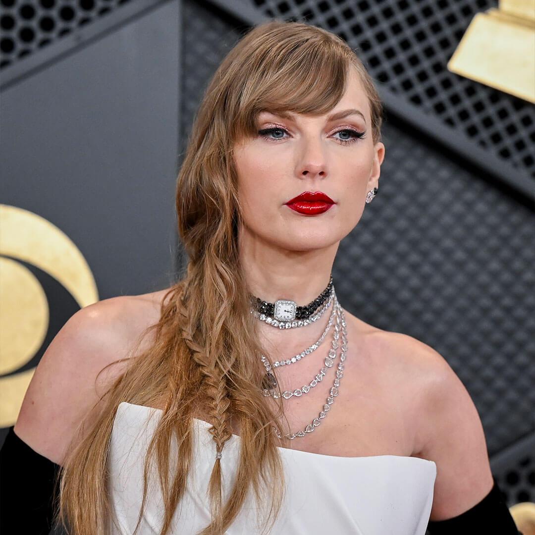 An image of Taylor Swift featuring her signature cat eyeliner and bold red lips, adorned with multiple gem necklaces, her hair is stylishly tucked down to the side, and she is wearing a white strapless dress along with black elbow-length gloves Desktop
