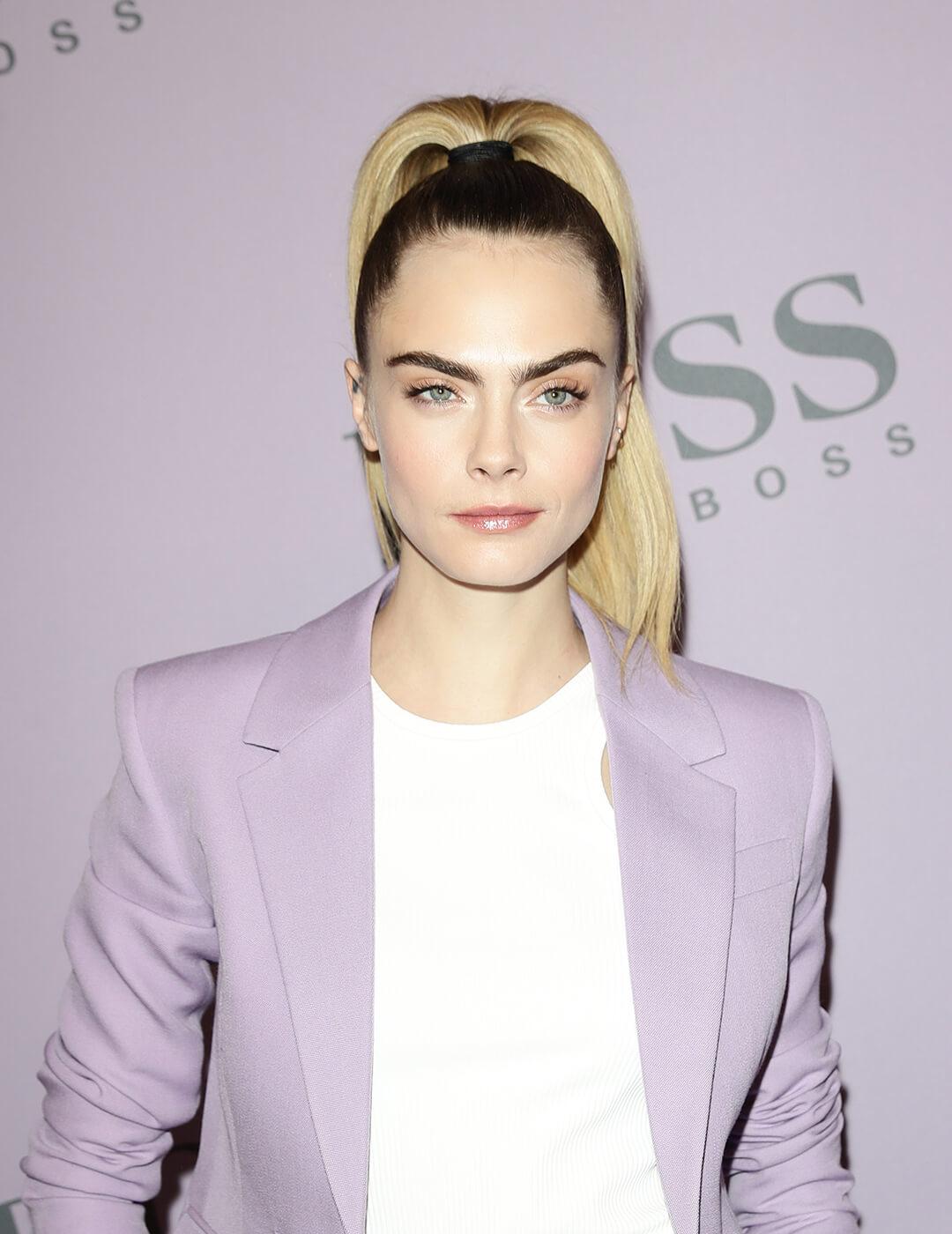 A photo of Cara Delevingne wearing a purple suit paired in white top with her blond hair styled in a simple classic chic ponytail Desktop