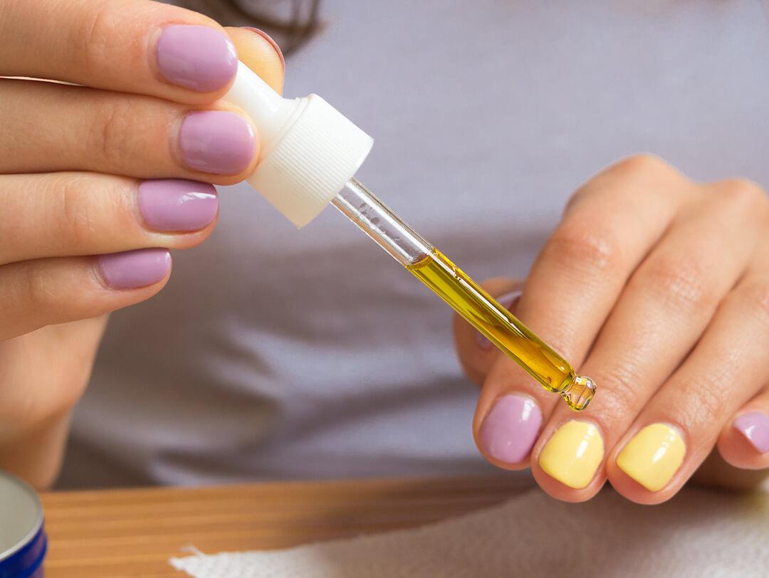 Close-up of a woman's hands with lavender and yellow nail polish applying oil on her cuticles with a dropper Desktop