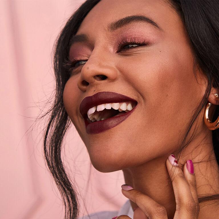 A closeup image of a model wearing rose gold eyeshadow and matte dark mauve lipstick and gold hoop earrings Desktop