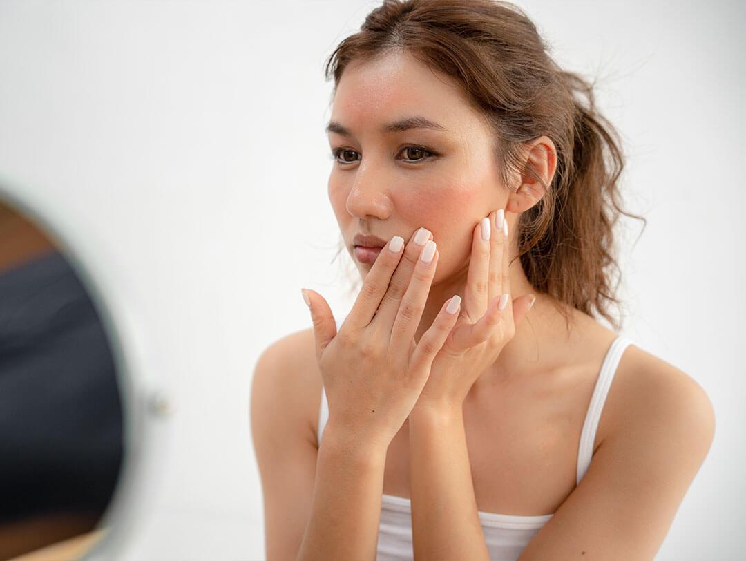 Woman with a thoughtful expression, touching her slightly reddened cheeks indicating inflammation, the soft lighting and natural background draw attention to her skin condition Desktop