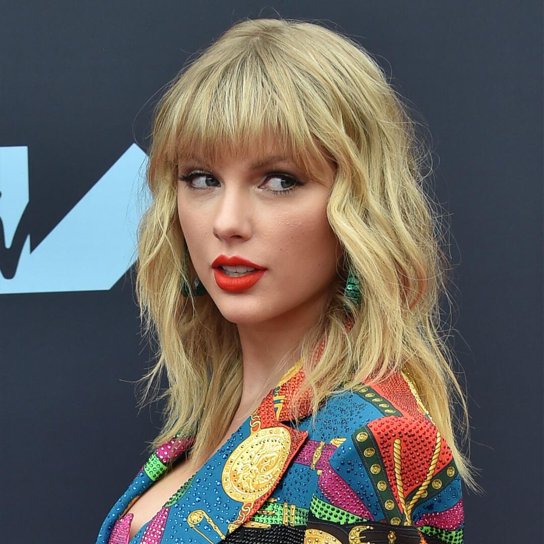 A picture of Taylor Swift wearing her signature pop of orange lipstick, gazing sideways with her wavy blonde hair cascading down Mobile