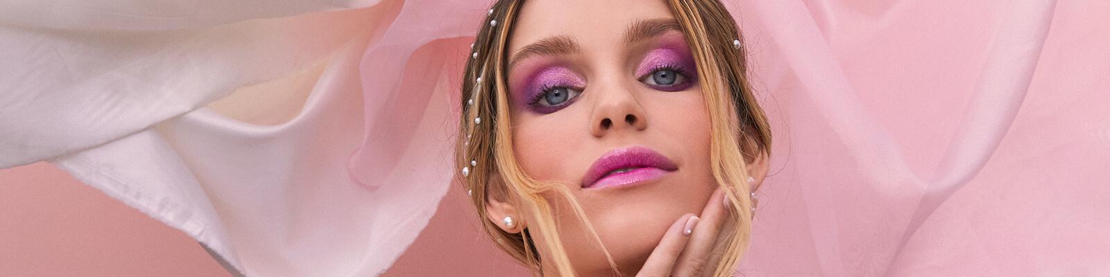 A closeup image of a model rocking a pink and purple smoky eyeshadow look paired with glossy pink lipstick feeling her jaw with one hand