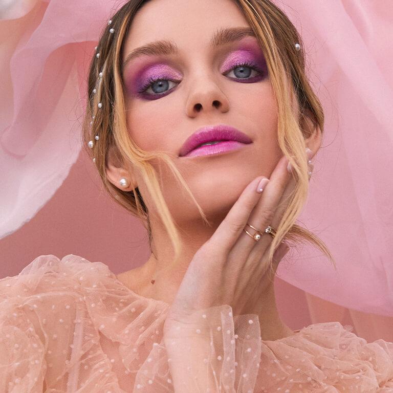 A closeup image of a model rocking a pink and purple smoky eyeshadow look paired with glossy pink lipstick feeling her jaw with one hand
