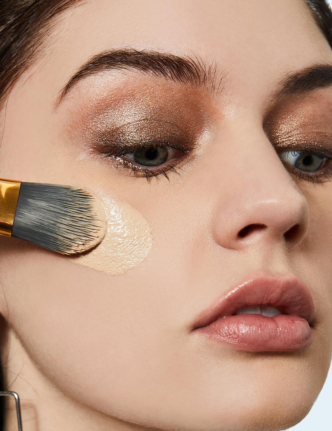 Close-up of a model applying applying foundation on her cheeks while wearing a shimmery nude eyeshadow Mobile