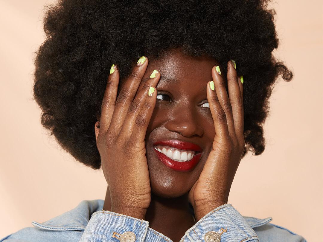 A photo of a model with curly hair touching her face with both hands on a light peach background Desktop