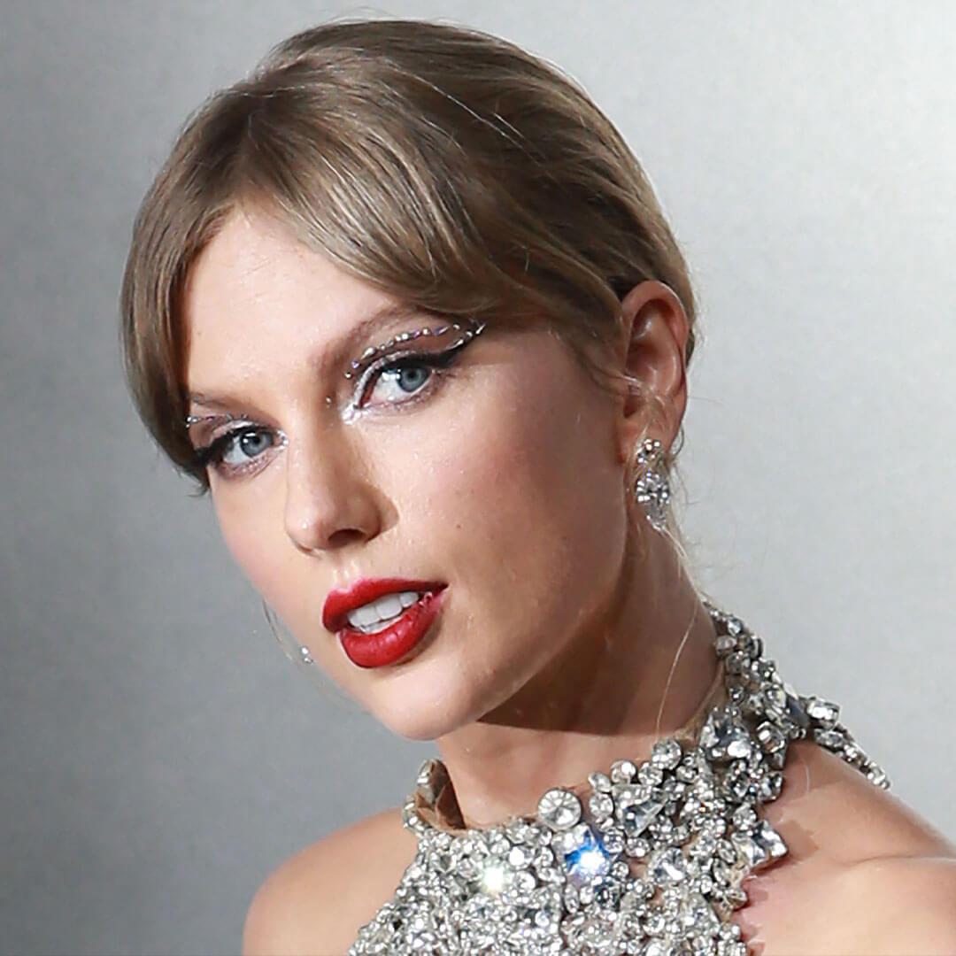 An image of Taylor Swift wearing a Bejeweled Sparkles see-through dress sheer dress made of nude mesh, featuring her bangs styled in a split and complemented by a striking red lipstick Mobile