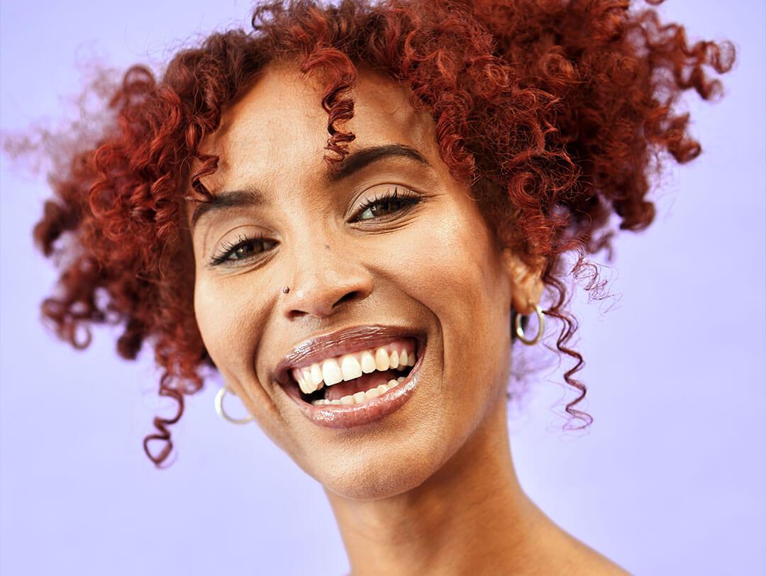 An image of a woman with color with a curly red hair styled in a two-piece messy bun wearing a bright blue spaghetti top while grinning at the camera Desktop