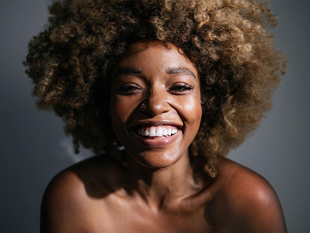 An image of a woman of color with an afro hairstyle is smiling at the camera against a gray backdrop Desktop
