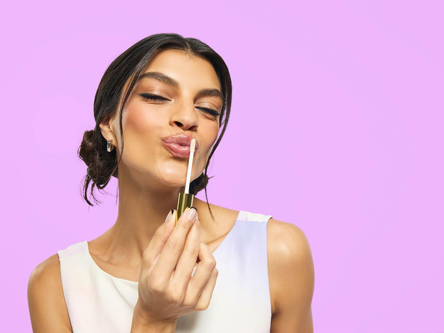 A woman pouts playfully while applying a shiny lip product with a gold applicator against a vibrant lavender background Desktop