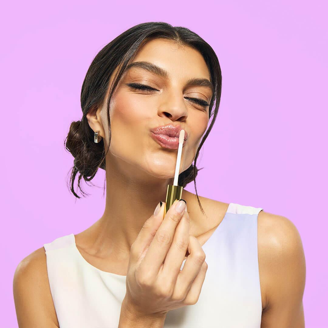 A woman pouts playfully while applying a shiny lip product with a gold applicator against a vibrant lavender background Mobile