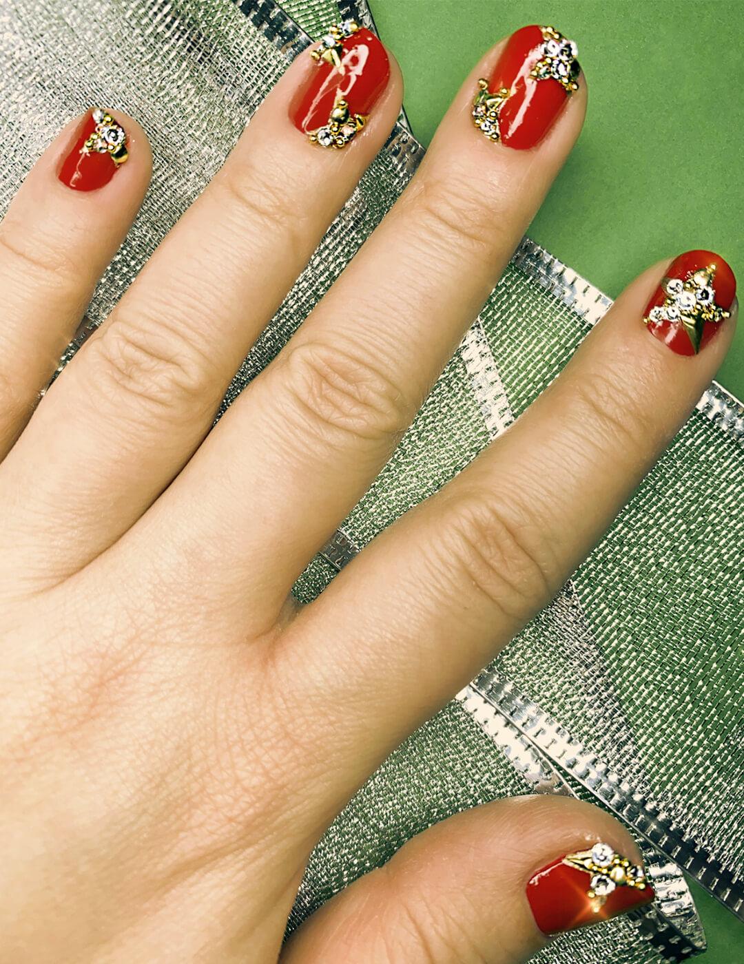 Close-up image of a model's hand with red mani embellished with rhinestones and studs against a green background with silver lace ribbons Desktop