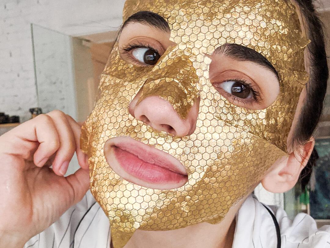 Photo of a young woman in her apartment putting on a gold sheet mask Desktop