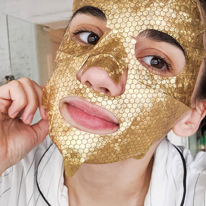 Photo of a young woman in her apartment putting on a gold sheet mask Mobile