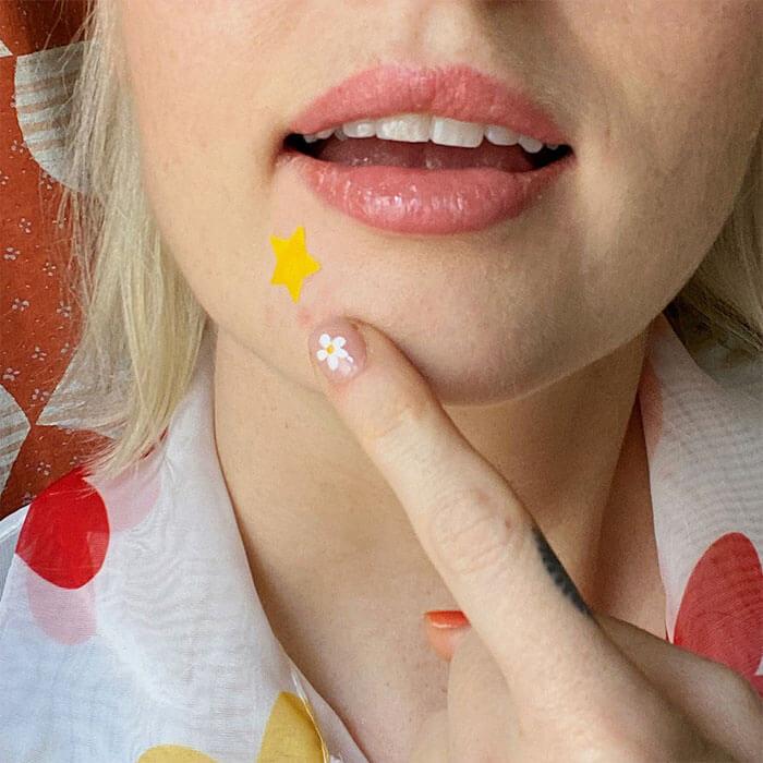 Woman with a star-shaped pimple patch on her face, touching it gently with a finger adorned with daisy nail art Mobile